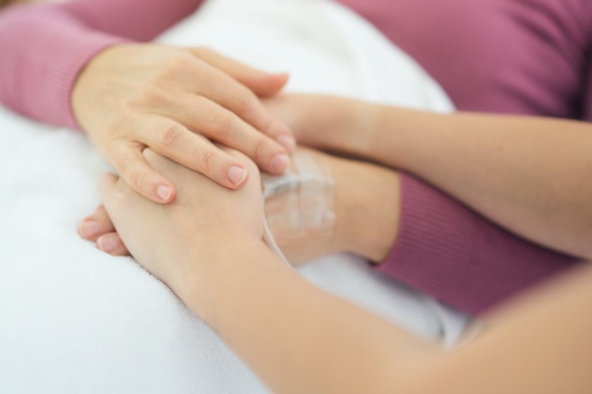 Dłoń bliskiej osoby spoczywa na ręce pacjenta z wkłuciem dożylnym, symbolizując wsparcie i troskę