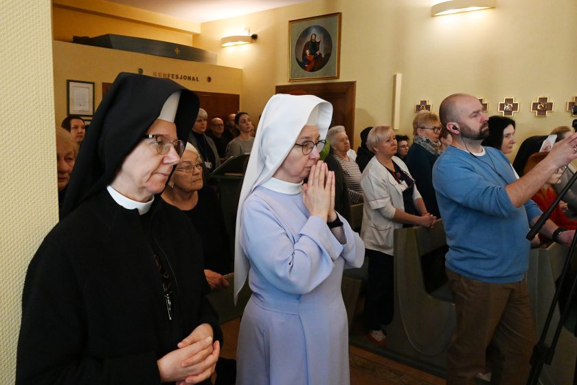 Siostry zakonne oraz wierni stoją w ławkach kaplicy podczas modlitwy; jedna z sióstr ma złożone dłonie, obok osoby świeckie skupione na liturgii