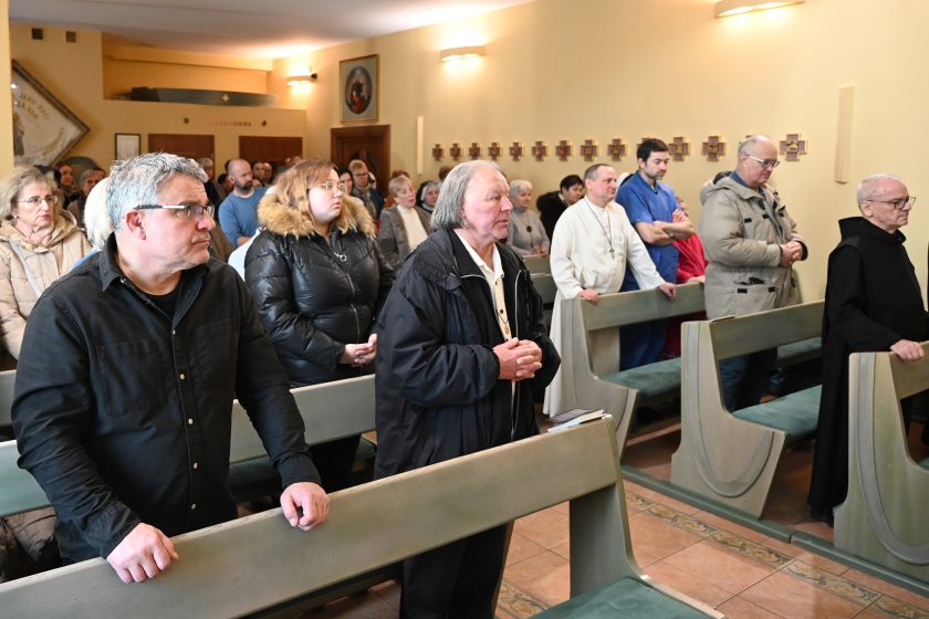 Wierni zgromadzeni w kaplicy stoją w ławkach podczas Mszy Świętej; na pierwszym planie mężczyzna z rękami opartymi na ławce, w tle liczna wspólnota uczestnicząca w modlitwie