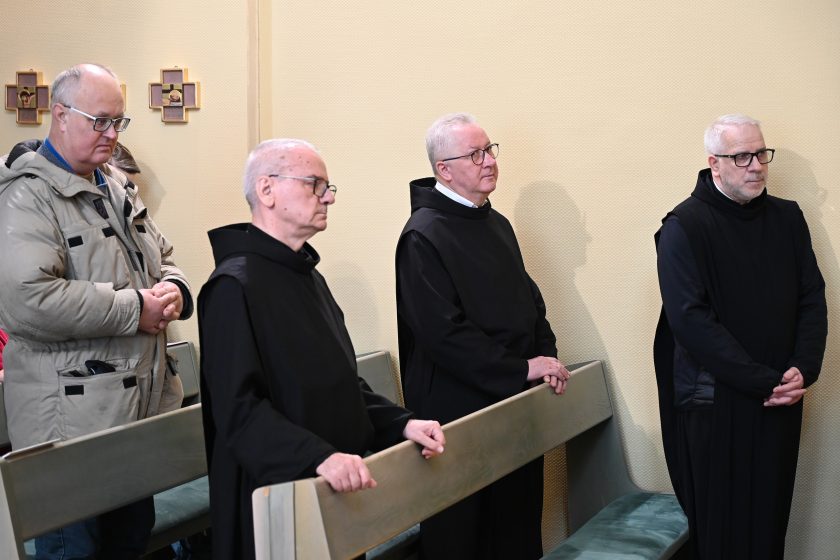 Bracia zakonni w czarnych habitach stoją w ławkach kaplicy podczas liturgii; obok nich świeccy uczestnicy Mszy Świętej
