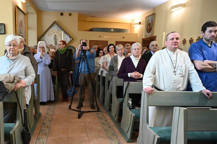 Widok na kaplicę wypełnioną wiernymi; część osób stoi w ławkach, w centralnej części ustawiony statyw z aparatem fotograficznym dokumentującym uroczystość