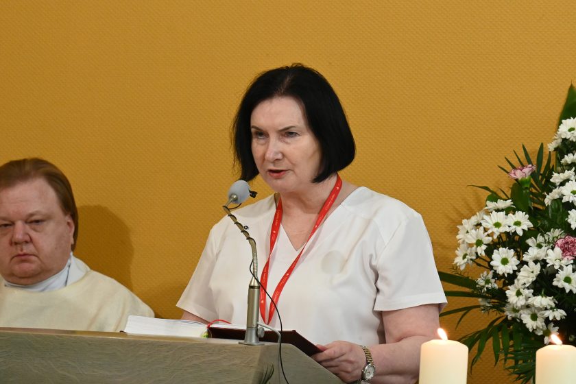 Kobieta w jasnym stroju medycznym z identyfikatorem na szyi czyta tekst z księgi przy ambonie; obok zapalona świeca i kompozycja z białych kwiatów, w tle siedzący duchowny