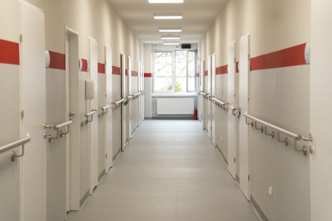 Długi, jasny korytarz szpitalny z białymi drzwiami po obu stronach. Na ścianach zamontowane są metalowe poręcze dla pacjentów oraz czerwony pas dekoracyjny biegnący wzdłuż korytarza. Na końcu znajduje się okno wpuszczające naturalne światło
