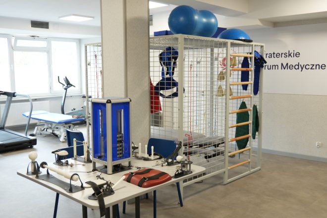 Sala rehabilitacyjna w Bonifraterskim Centrum Medycznym wyposażona w sprzęt do ćwiczeń i fizjoterapii. Widoczne są m.in. bieżnia, rower stacjonarny, stół do terapii, drabinki rehabilitacyjne oraz piłki gimnastyczne. Na ścianie znajduje się napis „Bonifraterskie Centrum Medyczne”