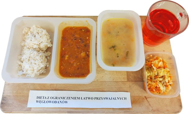 Obiad w diecie z ograniczeniem łatwo przyswajalnych węglowodanów podany na drewnianej desce. Na białej, plastikowej tacce znajduje się porcja ryżu oraz gulasz w czerwonym sosie warzywno-mięsnym. Obok ustawiony jest pojemnik z klarowną zupą jarzynową z widocznymi kawałkami warzyw oraz kubek z czerwonym napojem. Z prawej strony znajduje się osobny pojemnik z surówką z kapusty i marchwi. Przed zestawem leży kartka z napisem »DIETA Z OGRANICZENIEM ŁATWO PRZYSWAJALNYCH WĘGLOWODANÓW«