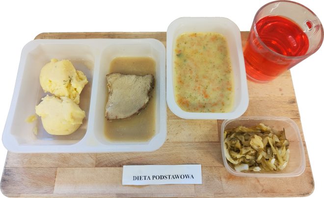 Obiad w diecie podstawowej podany na drewnianej tacy. Na białej, podzielonej tacce znajduje się porcja puree ziemniaczanego oraz plaster pieczonego mięsa podany z sosem. Obok umieszczono pojemnik z zupą jarzynową z widocznymi kawałkami warzyw oraz przezroczysty kubek z czerwonym napojem owocowym. Dodatkowo na tacy znajduje się osobny pojemnik z duszoną kapustą. Na środku tacy widoczna etykieta z napisem „Dieta podstawowa”