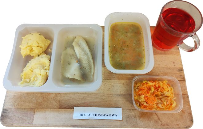 Obiad w diecie podstawowej podany na drewnianej tacy. W plastikowym dwudzielnym pojemniku znajduje się porcja puree ziemniaczanego oraz filet rybny w jasnym sosie. Obok stoi pojemnik z zupą jarzynową z widocznymi kawałkami warzyw i ziołami. Na tacy ustawiony jest szklany kubek z czerwonym napojem. W osobnym małym pojemniku znajduje się surówka z kiszonej kapusty i marchewki. Z przodu widoczna etykieta ‘DIETA PODSTAWOWA’