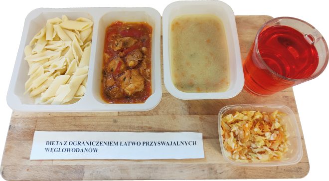 Obiad w diecie z ograniczeniem łatwo przyswajalnych węglowodanów, podany na drewnianej desce. Na białej, dzielonej tacce znajduje się porcja makaronu penne, mięso w sosie pomidorowym z warzywami oraz zupa jarzynowa. Obok stoi szklanka czerwonego napoju. Z boku widoczna jest osobna porcja surówki z kapusty i marchwi. Na desce umieszczona kartka z napisem: ‘Dieta z ograniczeniem łatwo przyswajalnych węglowodanów