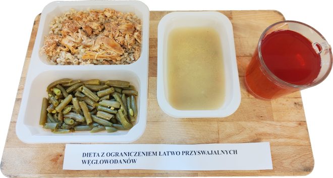 Obiad w ramach diety z ograniczeniem łatwo przyswajalnych węglowodanów: kasza z kawałkami gotowanego mięsa drobiowego, fasolka szparagowa oraz porcja bulionu w osobnym pojemniku. Obok szklanka czerwonego napoju. Na stole kartka z napisem „Dieta z ograniczeniem łatwo przyswajalnych węglowodanów”