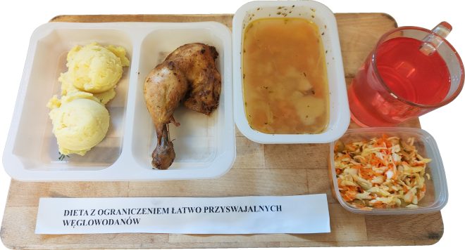 Na drewnianej desce zaprezentowano zestaw obiadowy z kartką z napisem „DIETA Z OGRANICZENIEM ŁATWO PRZYSWAJALNYCH WĘGLOWODANÓW”. W plastikowych pojemnikach znajdują się: pieczona ćwiartka kurczaka oraz dwie porcje puree ziemniaczanego uformowane w kulki. W osobnym pojemniku podano klarowną zupę z widocznymi kawałkami warzyw. Obok ustawiono pojemnik z surówką z kapusty białej, marchwi i ogórka kiszonego. Całość uzupełnia szklany kubek z czerwonym napojem, prawdopodobnie herbatą owocową