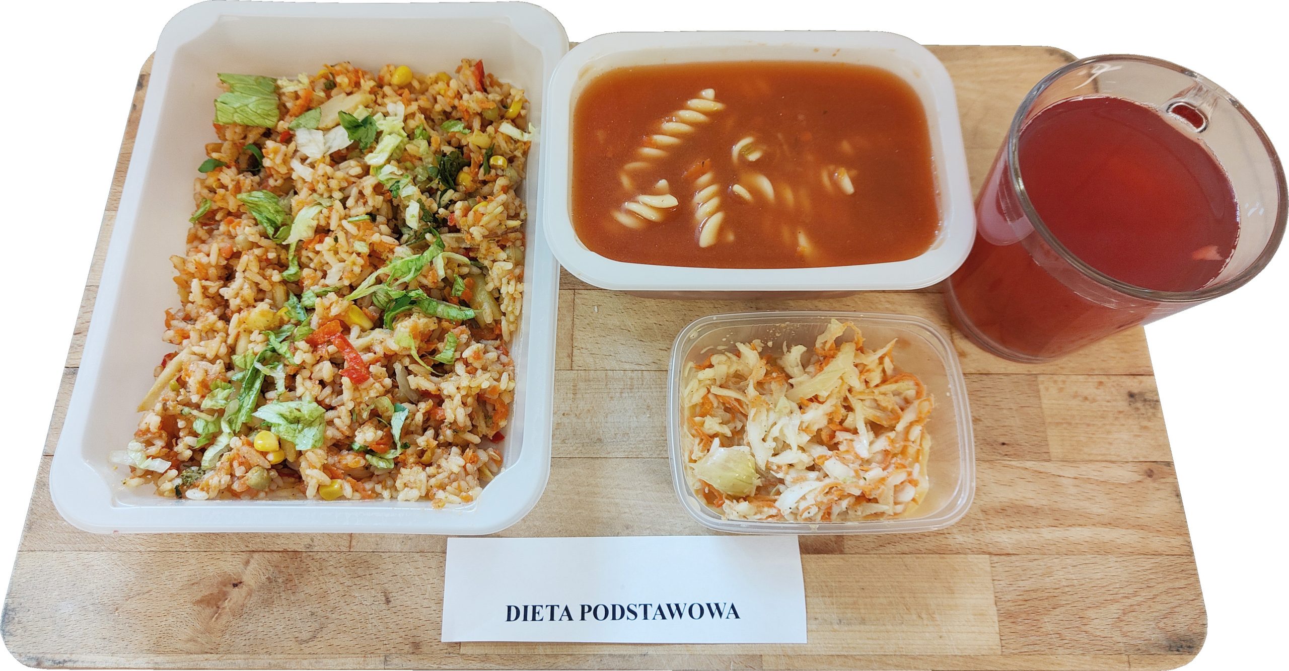Na drewnianej desce ustawiono zestaw obiadowy w białych pojemnikach. Po lewej znajduje się duża porcja ryżu wymieszanego z warzywami (widać m.in. kukurydzę i kawałki papryki) oraz poszarpaną sałatą i zieleniną na wierzchu. Pośrodku w prostokątnym pojemniku jest czerwona zupa z makaronem w kształcie świderków. Z przodu stoi mały, przezroczysty pojemnik z surówką z poszatkowanej kapusty i marchewki. Po prawej umieszczono szklany kubek z czerwonym napojem. Na dole kadru leży kartka z napisem „DIETA PODSTAWOWA”