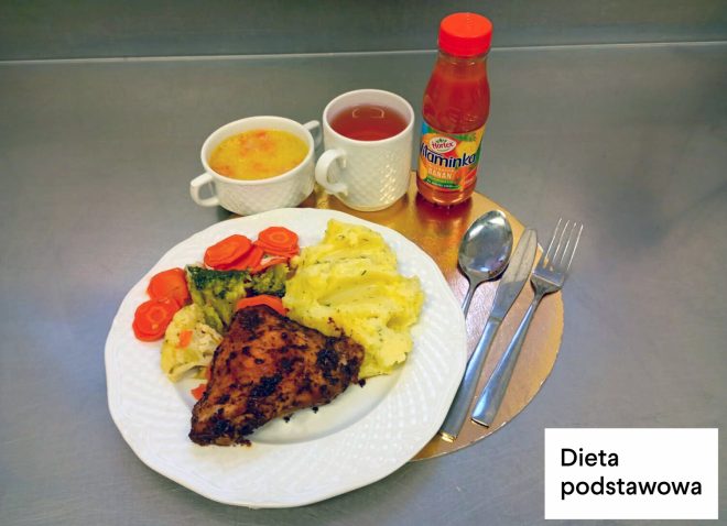 Obiad w diecie podstawowej: na talerzu pieczone udko z kurczaka, gotowane warzywa – marchew i brokuły – oraz ziemniaki z koperkiem. Obok stoją kubek z zupą, kubek z herbatą oraz butelka soku. W prawym dolnym rogu napis „Dieta podstawowa”