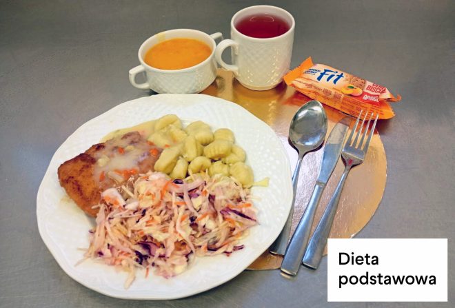 Obiad w diecie podstawowej: na talerzu kotlet z sosem, kopytka oraz surówka z białej i czerwonej kapusty. Obok stoją dwa kubki z zupą i napojem oraz zapakowany baton. W prawym dolnym rogu napis „Dieta podstawowa”