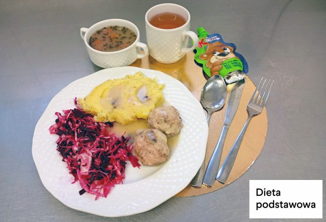 Obiad w diecie podstawowej: na talerzu puree ziemniaczane z sosem, dwa pulpety oraz surówka z buraczków i kapusty. Obok znajduje się kubek z zupą, kubek z herbatą oraz mus owocowy w opakowaniu. W prawym dolnym rogu napis „Dieta podstawowa”