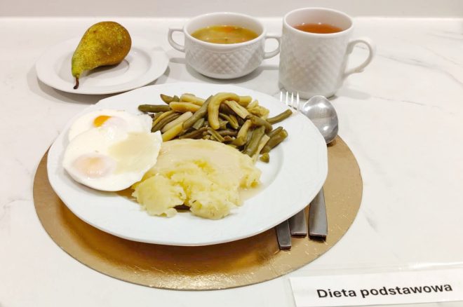 Obiad w diecie podstawowej: na białym talerzu puree ziemniaczane, jajka sadzone oraz gotowana fasolka szparagowa. W tle gruszka na talerzyku, miseczka z zupą jarzynową oraz kubek z herbatą. Zestaw podany na tacy