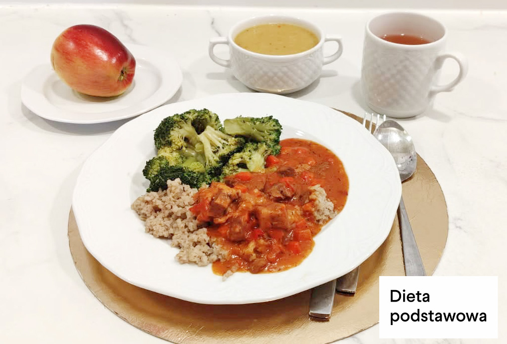 Obiad w diecie podstawowej podany na dużym białym talerzu. Na talerzu znajduje się porcja kaszy, gotowane brokuły oraz mięso w sosie pomidorowym z kawałkami warzyw. W tle stoją dwa białe naczynia – miseczka z zupą krem oraz kubek z herbatą. Po lewej stronie na małym talerzyku leży czerwone jabłko. W prawym dolnym rogu zdjęcia widoczny napis „Dieta podstawowa”