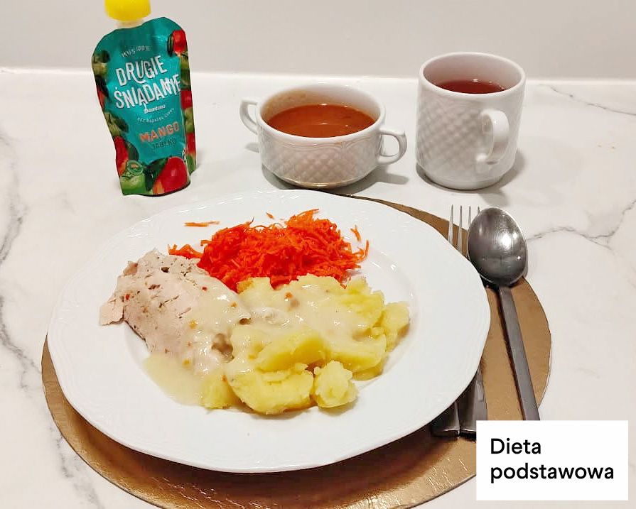 Na talerzu gotowany filet drobiowy polany jasnym sosem, puree ziemniaczane oraz surówka z tartej marchewki. W tle kubek herbaty, filiżanka zupy oraz mus owocowy w tubce. Danie podane na białej zastawie ze sztućcami po prawej stronie