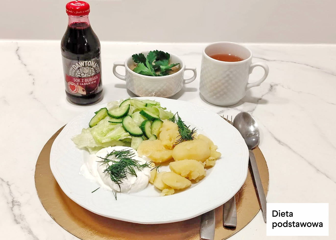 Na białym talerzu jajko sadzone z koperkiem, gotowane ziemniaki posypane koperkiem oraz sałatka z ogórka i sałaty. W tle kubek herbaty, filiżanka zupy oraz butelka soku z buraka i jabłka. Sztućce ułożone obok talerza