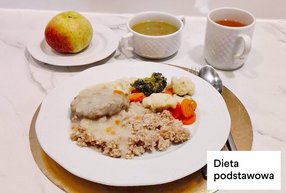 Kasza z sosem i dwa kawałki mięsa w jasnym sosie, podane z gotowanymi warzywami: kalafiorem, brokułem i marchewką. Obok talerza jabłko na osobnym spodku, w tle filiżanka zupy i kubek herbaty