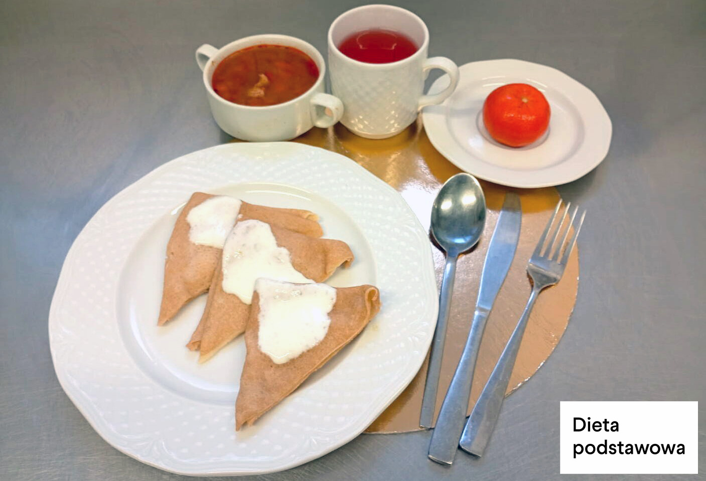 Trzy naleśniki z jasnym sosem podane na białym talerzu. W tle filiżanka zupy, kubek herbaty oraz mandarynka na małym talerzyku. Sztućce ułożone po prawej stronie