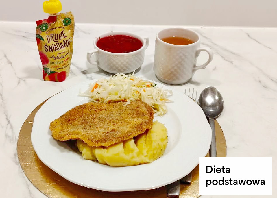 Kotlet schabowy w panierce podany z ziemniakami puree oraz surówką z białej kapusty. W tle filiżanka czerwonej zupy, kubek herbaty oraz mus owocowy w tubce. Danie ustawione na złotej podkładce