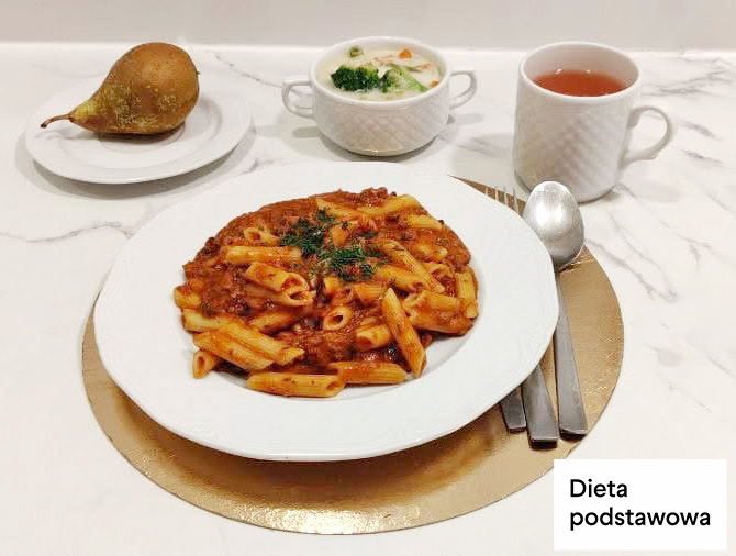 Talerz z makaronem penne w sosie pomidorowym, posypanym natką pietruszki. Obok miseczka z zupą jarzynową, kubek herbaty oraz gruszka na osobnym talerzyku. Dieta podstawowa