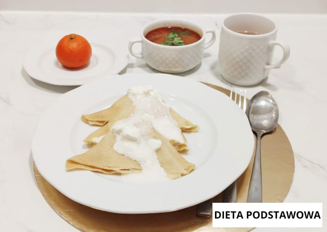 Posiłek w diecie podstawowej podany na dużym białym talerzu. Na środku znajdują się trzy złożone naleśniki polane białym sosem. W tle ustawiono mały talerzyk z mandarynką, miskę z czerwonym barszczem z natką pietruszki oraz kubek z herbatą. Po prawej stronie widoczne są sztućce: łyżka i widelec. W prawym dolnym rogu znajduje się napis ‘DIETA PODSTAWOWA’