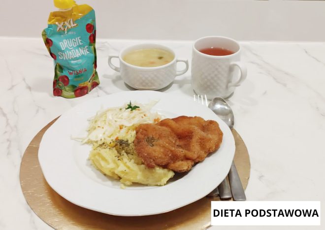 Obiad w diecie podstawowej podany na białym talerzu na złotej podkładce. Na talerzu znajduje się panierowany kotlet, porcja puree ziemniaczanego oraz surówka z białej kapusty. W tle stoją dwa naczynia: miseczka z jasną zupą oraz kubek z herbatą. Po lewej stronie widoczna jest tubka musu owocowego z napisem ‘Drugie śniadanie, wiśnia’. W prawym dolnym rogu znajduje się napis ‘DIETA PODSTAWOWA’