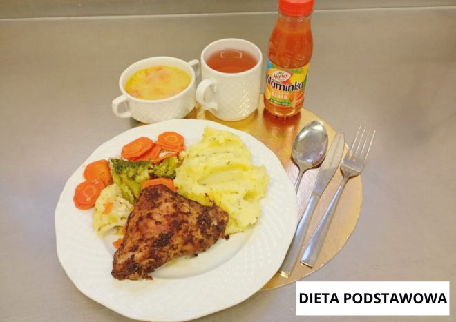 Obiad w diecie podstawowej: na białym talerzu porcja puree ziemniaczanego, pieczony filet drobiowy oraz gotowane warzywa – marchew i brokuł. W tle miseczka z zupą jarzynową, kubek z herbatą oraz butelka napoju owocowego. Sztućce ułożone po prawej stronie talerza