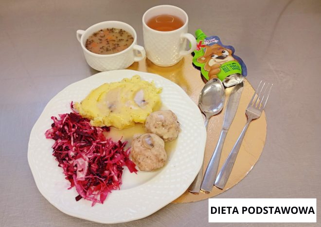 Obiad w diecie podstawowej: na białym talerzu puree ziemniaczane polane sosem, dwa pulpety oraz surówka z buraków. W tle miseczka z zupą, kubek z herbatą oraz opakowanie musu owocowego. Sztućce ułożone obok talerza