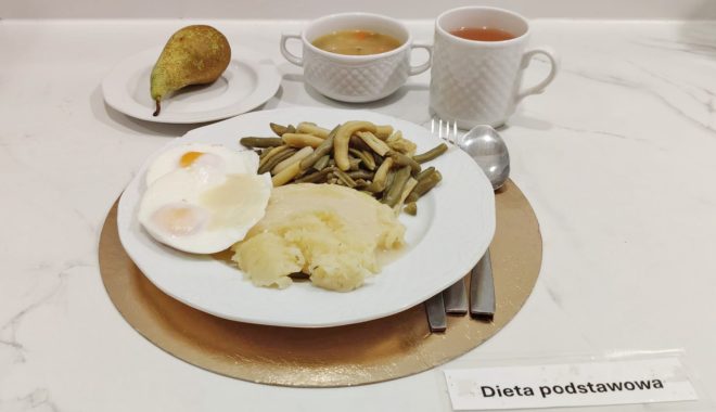 Obiad w ramach diety podstawowej: ziemniaki, jajka sadzone oraz fasolka szparagowa na białym talerzu. W tle gruszka na osobnym talerzyku, miseczka z zupą oraz kubek z herbatą. Na stole znajduje się etykieta „Dieta podstawowa” oraz sztućce