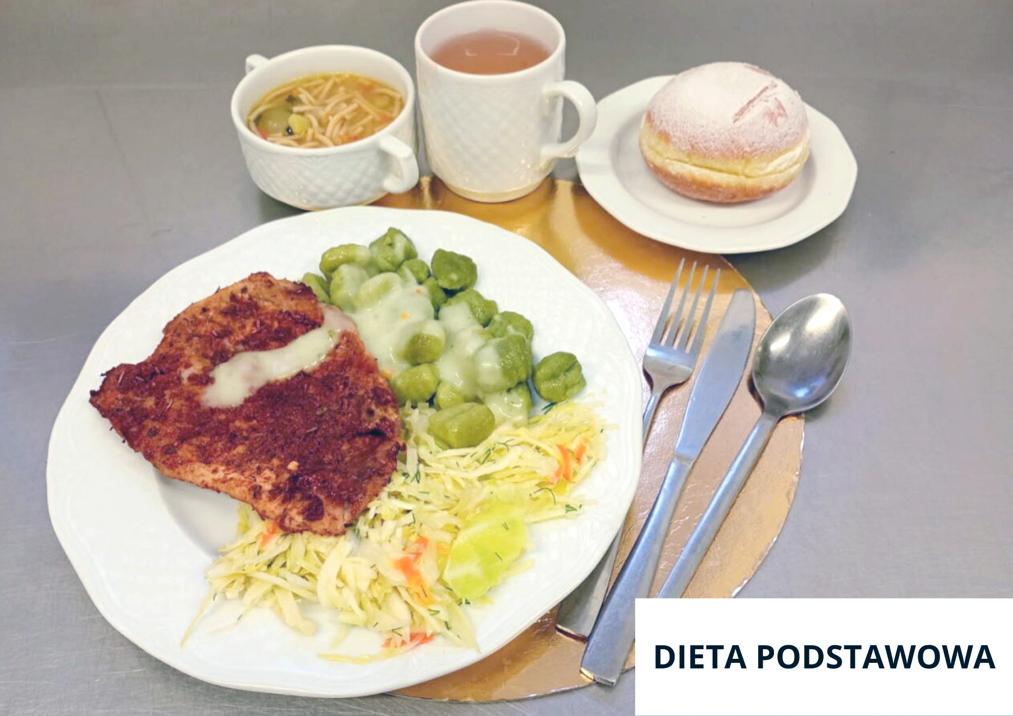 Na stalowym blacie na złotej podkładce ustawiono zestaw obiadowy. Na dużym białym talerzu znajduje się panierowany, rumiany kotlet polany jasnym sosem. Obok kotleta leżą kluski śląskie w jasnym sosie oraz porcja surówki z jasnej kapusty z dodatkiem koperku i kawałków marchewki. Z tyłu stoją dwie białe filiżanki: w jednej zupa z cienkim makaronem i widocznymi warzywami, w drugiej jasnoróżowy napój (kompot lub herbata). Po prawej stronie na małym talerzyku leży pączek posypany cukrem pudrem. Z prawej strony podkładki ułożone są sztućce: widelec, nóż i łyżka. W prawym dolnym rogu widoczny napis „DIETA PODSTAWOWA”