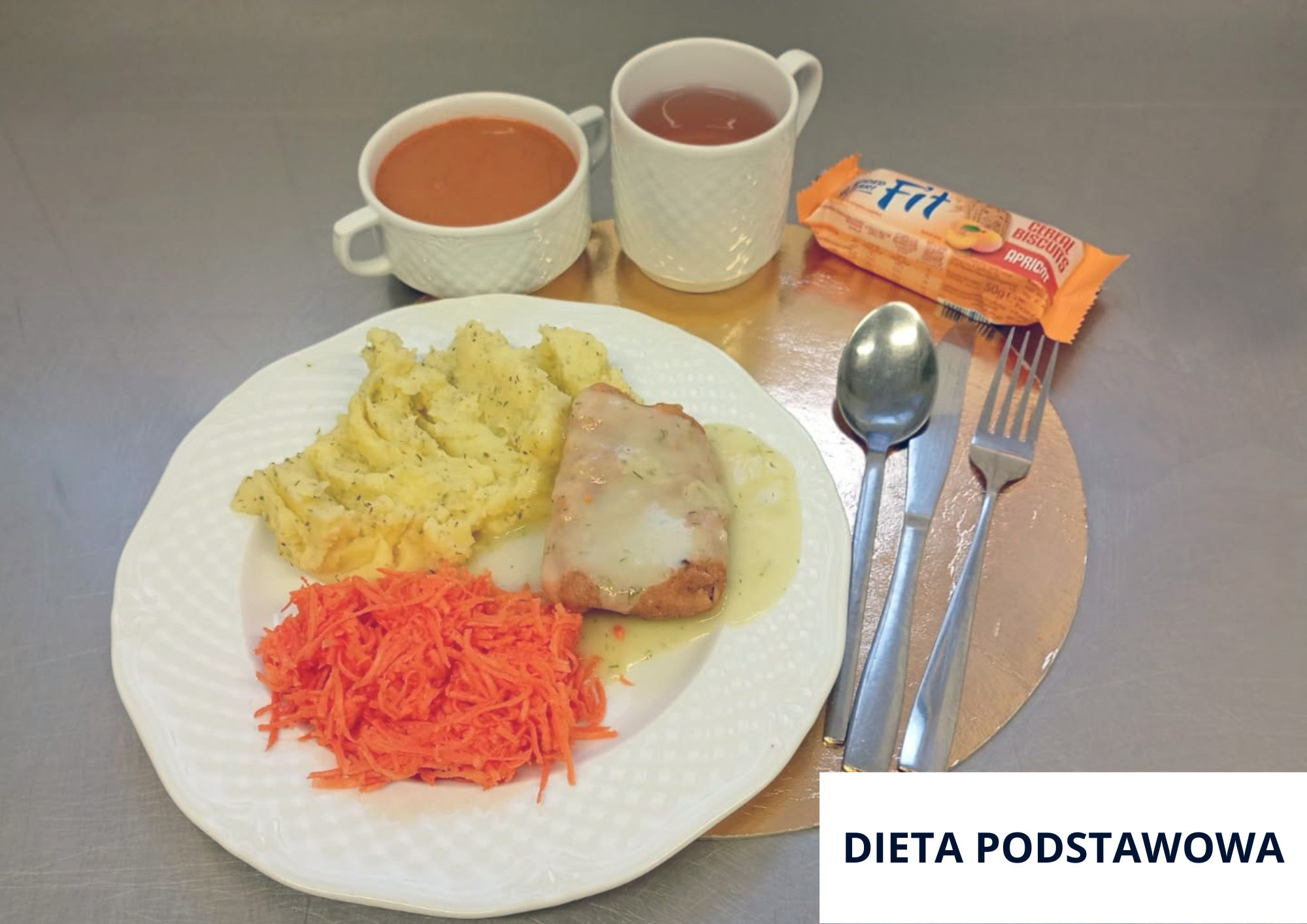 Na stalowym blacie na złotej podkładce stoi biały talerz z porcją ziemniaków puree z zielonymi ziołami. Obok puree znajduje się kawałek mięsa w jasnym sosie oraz porcja surówki z tartej marchewki. Z tyłu ustawiono dwie białe filiżanki: w jednej czerwony napój (kompot/herbata), w drugiej zupa-krem w odcieniu pomarańczowym. Po prawej stronie, na podkładce, leży zapakowany baton/ciastka „Fit” o smaku morelowym. Obok znajdują się sztućce: łyżka, nóż i widelec. W prawym dolnym rogu znajduje się biała plansza z napisem „DIETA PODSTAWOWA”