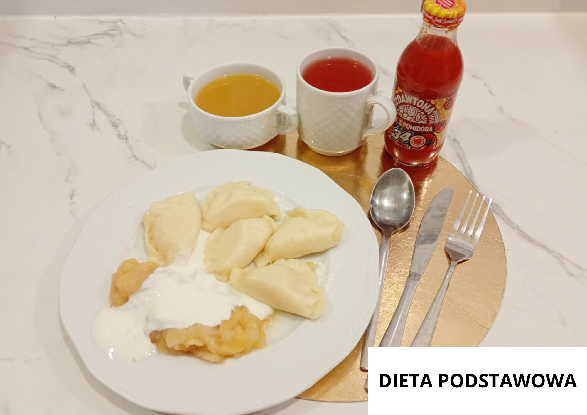 Na jasnym, marmurkowym blacie na złotej podkładce stoi biały talerz z pierogami polanymi białym sosem (śmietanowym). Obok pierogów widoczna jest porcja musu lub puree (jasnobeżowego) również częściowo przykryta sosem. Z tyłu ustawiono dwa naczynia: białą filiżankę z żółtą zupą/bulionem oraz biały kubek z czerwonym napojem (kompot). Po prawej stronie stoi butelka czerwonego soku/napoju pomidorowego z etykietą. Po prawej stronie talerza leżą sztućce: łyżka, nóż i widelec. W prawym dolnym rogu widoczny napis „DIETA PODSTAWOWA”