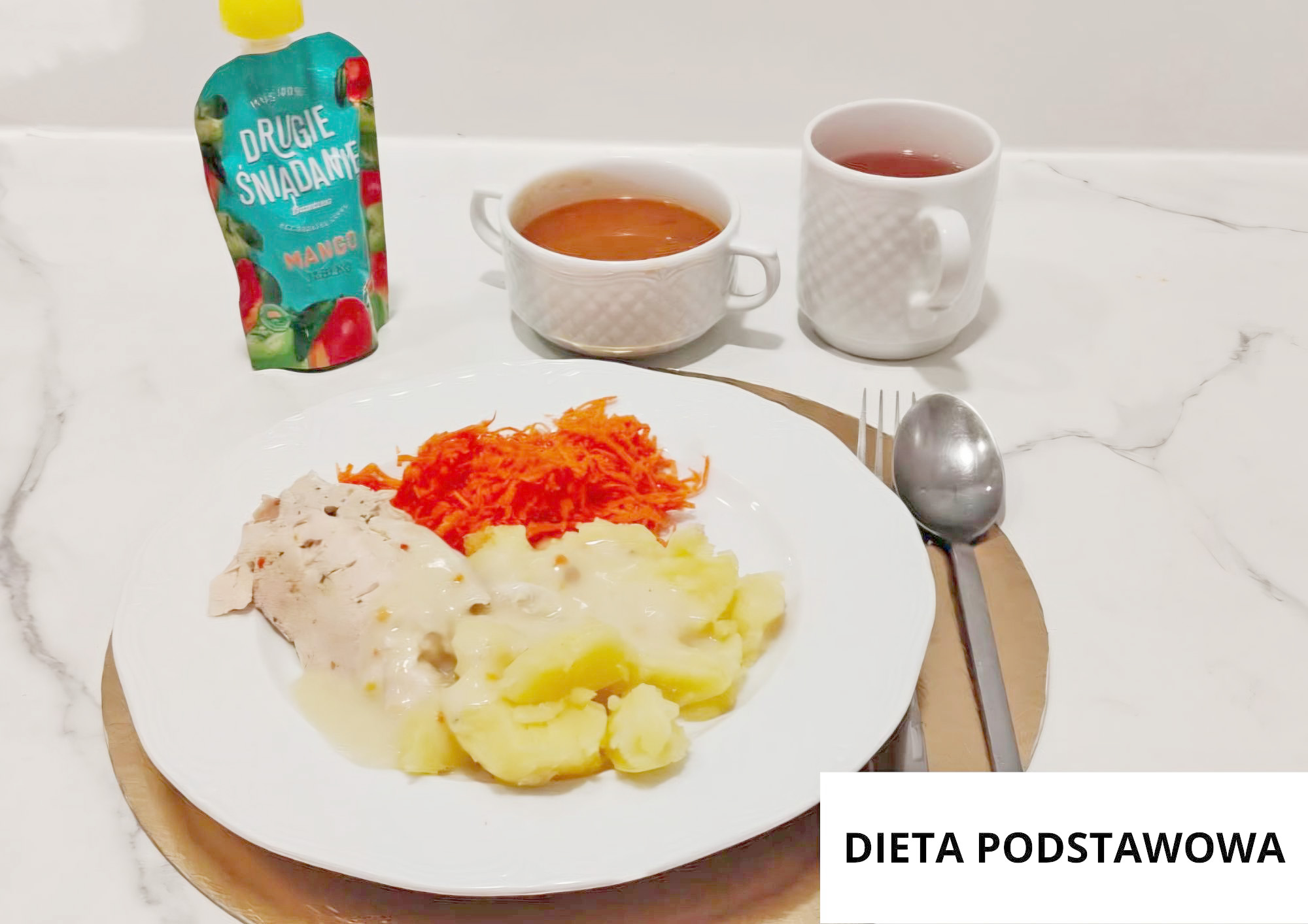 Na jasnym, marmurkowym blacie na złotej podkładce stoi biały talerz obiadowy. Na talerzu ułożono gotowane ziemniaki polane jasnym sosem oraz plaster białego mięsa (np. drobiu) również w sosie. Z tyłu znajduje się porcja tartej marchewki jako surówka, a obok stoją dwa naczynia: biała filiżanka z pomarańczową zupą-krem oraz biały kubek z herbatą lub kompotem. Po lewej stronie zestawu stoi saszetka z napisem „DRUGIE ŚNIADANIE” (smak mango) w kolorowej oprawie. Po prawej stronie leżą sztućce: widelec i łyżka. W prawym dolnym rogu widoczna biała plansza z napisem „DIETA PODSTAWOWA”