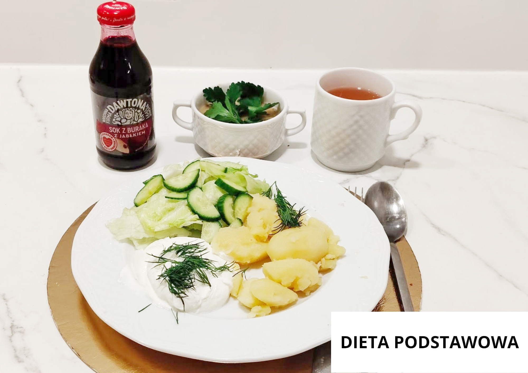 Na białym talerzu jajko sadzone posypane koperkiem, gotowane ziemniaki z koperkiem oraz sałatka z plasterków ogórka i liści sałaty. W tle filiżanka zupy, kubek herbaty oraz butelka soku z buraka i jabłka. Sztućce ułożone po prawej stronie talerza, całość na złotej podkładce
