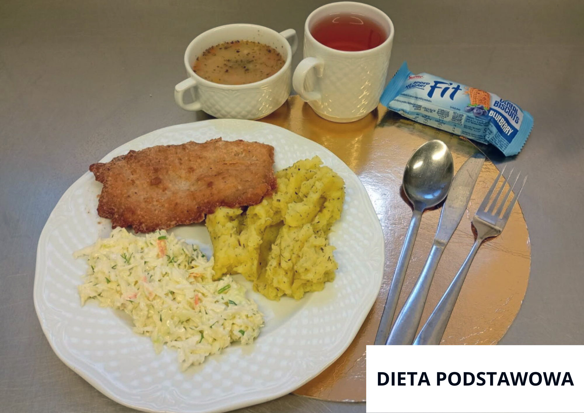 Kotlet schabowy w złocistej panierce podany z puree ziemniaczanym z ziołami oraz surówką z białej kapusty z koperkiem i marchewką. W tle filiżanka zupy, kubek herbaty oraz opakowanie herbatników. Nakrycie ustawione na złotej podkładce, sztućce po prawej stronie