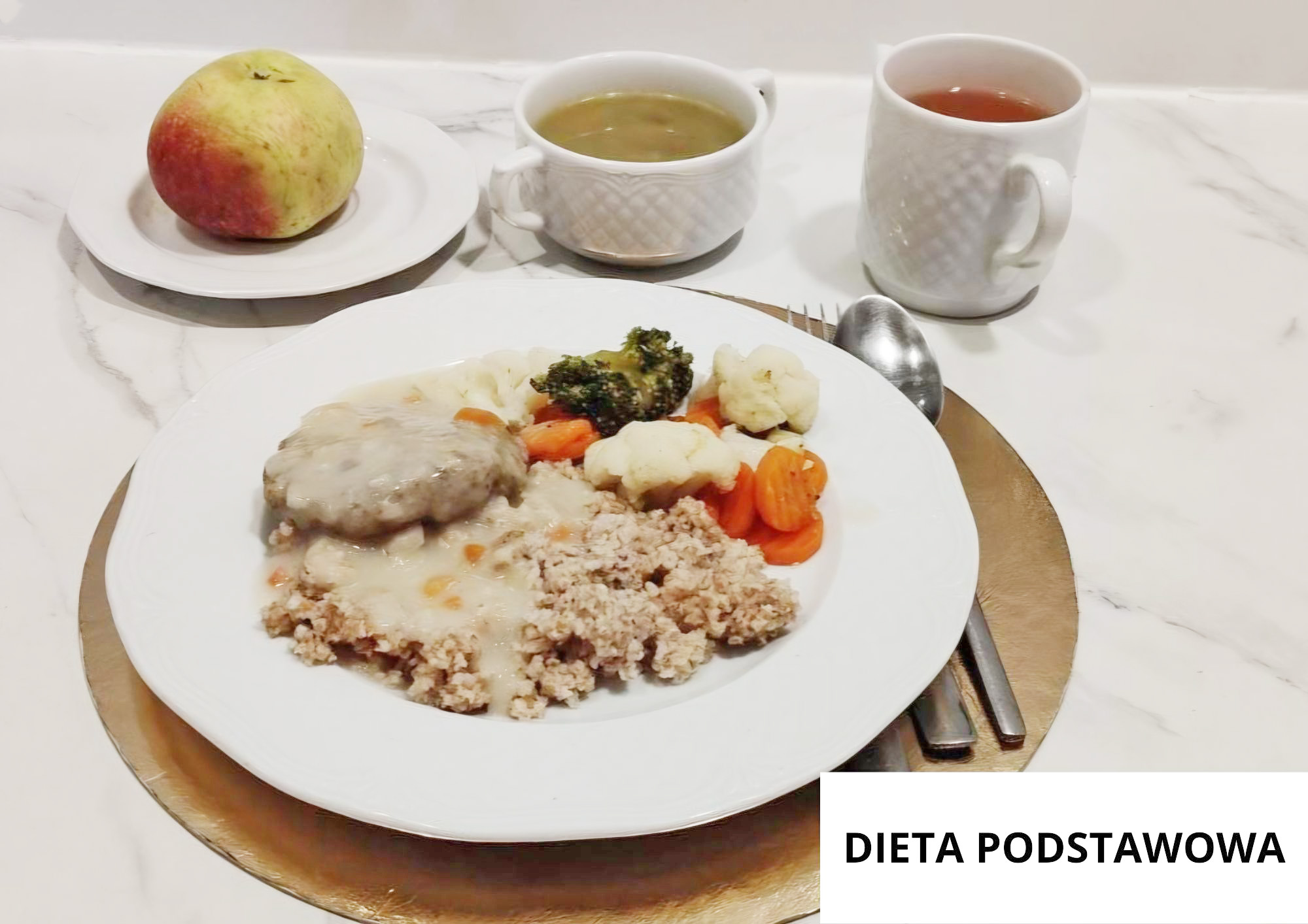Kasza z sosem oraz dwa kawałki mięsa w jasnym sosie, podane z gotowanymi warzywami: kalafiorem, brokułem i marchewką. Obok na osobnym talerzyku jabłko. W tle filiżanka zupy i kubek herbaty. Talerz ustawiony na złotej podkładce