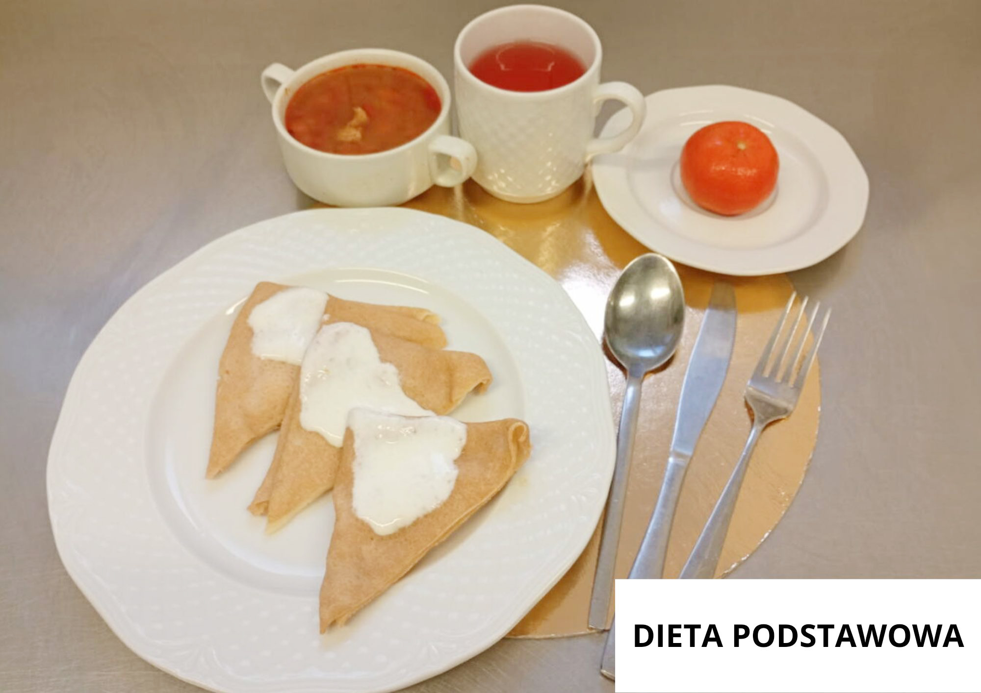 Trzy naleśniki z jasnym sosem podane na białym talerzu. W tle filiżanka zupy pomidorowej, kubek herbaty oraz mandarynka na małym talerzyku. Sztućce ułożone po prawej stronie, całość na złotej podkładce