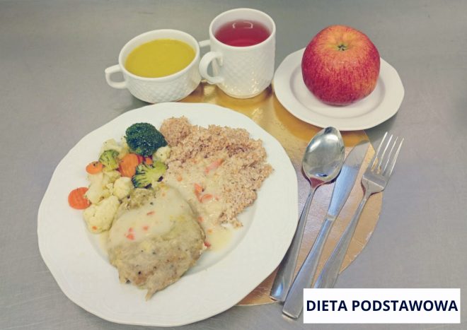 „Obiad w diecie podstawowej podany na białym talerzu ustawionym na złotej podkładce. Na talerzu znajduje się porcja kaszy, filet drobiowy w jasnym sosie oraz gotowane warzywa: brokuły, kalafior i marchew. W tle stoją dwa naczynia: miseczka z żółtą zupą oraz kubek z czerwonym napojem. Obok na małym talerzyku leży czerwone jabłko. Po prawej stronie ułożone są sztućce. W prawym dolnym rogu widoczny napis ‘DIETA PODSTAWOWA