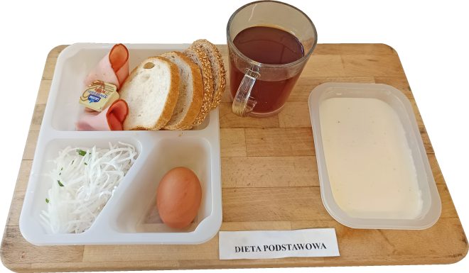 Śniadanie w diecie podstawowej podane na drewnianej desce. Na białej, plastikowej tacce znajdują się kromki pieczywa pszennego i z ziarnami, plasterki wędliny drobiowej, mały pakowany serek topiony, tarta biała rzodkiewka oraz jajko gotowane na twardo. Obok tacy stoi szklany kubek z herbatą oraz zamknięty pojemnik z jasnym jogurtem lub kefirem. Przed tacą widoczna kartka z napisem »DIETA PODSTAWOWA«