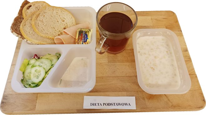 Śniadanie w diecie podstawowej podane na drewnianej tacy. Na białej, podzielonej tacce znajdują się kromki pieczywa pszennego i razowego, plastry wędliny drobiowej oraz mała porcja masła w jednorazowym opakowaniu. W osobnych przegródkach podano sałatkę z liści sałaty i plasterków świeżego ogórka oraz jasny sos jogurtowy. Obok znajduje się przezroczysty kubek z herbatą oraz pojemnik z jasną owsianką na mleku. Na tacy widoczna etykieta z napisem „Dieta podstawowa”