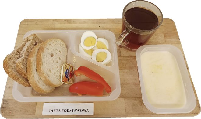 Śniadanie w diecie podstawowej podane na drewnianej tacy. W plastikowym pojemniku znajdują się kromki chleba mieszanego i pszennego, porcje masła oraz plasterki czerwonej papryki. Obok ułożone są kawałki jajka na twardo. Na tacy stoi szklany kubek z herbatą, a w osobnym pojemniku znajduje się jasna zupa mleczna. Z przodu widoczna etykieta z napisem ‘DIETA PODSTAWOWA’