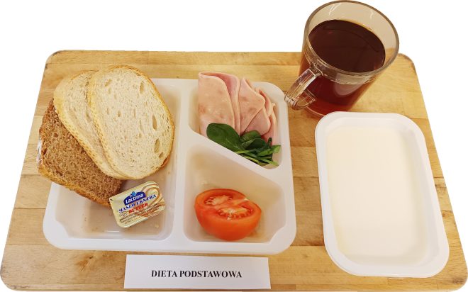 Śniadanie w ramach diety podstawowej: kromki chleba pszennego i pełnoziarnistego, masło porcjowane, plasterki wędliny drobiowej, pomidor oraz świeże liście szpinaku. Obok kubek z herbatą i dodatkowy pojemnik. Na stole widoczna kartka z napisem „Dieta podstawowa”