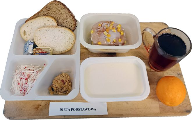 Na drewnianej desce ustawiono zestaw posiłków oznaczony kartką z napisem „DIETA PODSTAWOWA”. W plastikowych, białych pojemnikach znajdują się: dwie kromki chleba pszennego i jedna kromka chleba razowego, porcja masła w kostce oraz serek topiony w trójkątnym opakowaniu. Obok widoczna jest surówka z białej rzodkwi z dodatkiem drobnych czerwonych kawałków oraz porcja pasty kanapkowej. W osobnym pojemniku znajduje się jasna zupa krem. Na deser podano drożdżówkę z lukrem i kandyzowaną skórką pomarańczową. Obok stoi szklany kubek z herbatą oraz świeża pomarańcza