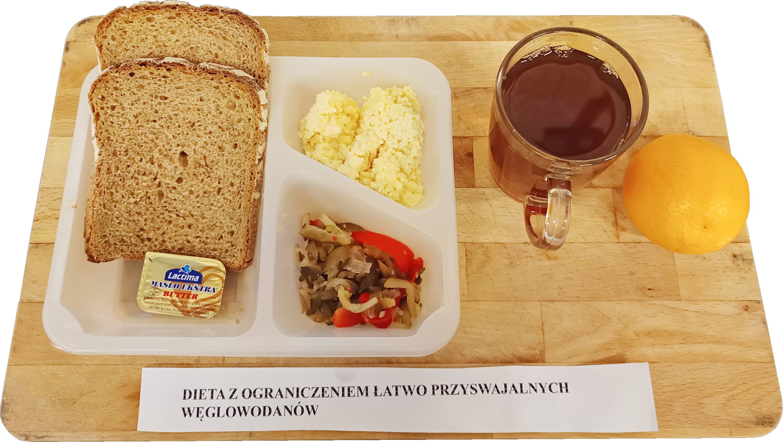 Taca z posiłkiem oznaczonym „Dieta z ograniczeniem łatwo przyswajalnych węglowodanów”. Na białej, trójdzielnej tacce znajdują się dwie kromki chleba razowego, porcja twarogu, surówka z pieczarek i czerwonej papryki oraz kostka masła Lactima. Obok stoi przezroczysty kubek z herbatą oraz leży cały grejpfrut. Całość ustawiona na jasnym drewnianym blacie