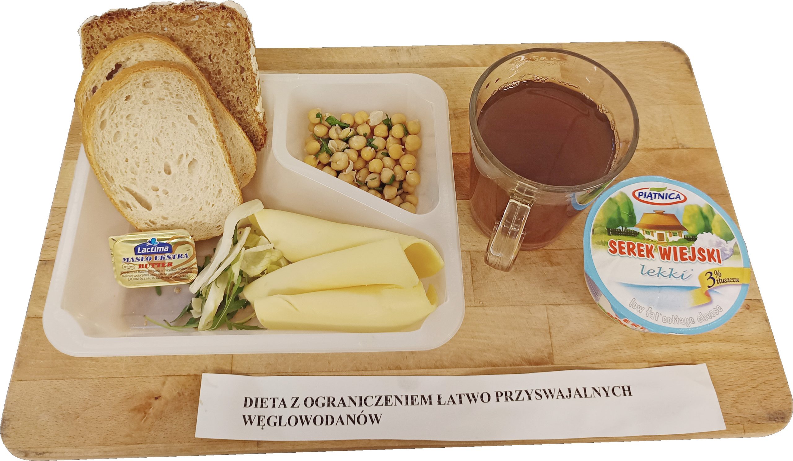 Na drewnianej desce podano śniadanie ułożone na białej tacy z przegródkami. Po lewej stronie leżą kromki pieczywa: jasne i ciemniejsze, pełnoziarniste. Na tacy widać małą porcję masła w złotym opakowaniu, kilka zwiniętych plastrów żółtego sera oraz niewielką porcję posiekanej zieleniny. W górnej przegródce znajduje się sałatka z ziaren ciecierzycy z dodatkiem drobno posiekanych składników. Po prawej stronie stoi szklany kubek z herbatą, a obok zamknięte opakowanie „Serek wiejski lekki” (Piątnica). Na dole kadru widoczna kartka z napisem „DIETA Z OGRANICZENIEM ŁATWO PRZYSWAJALNYCH WĘGLOWODANÓW”