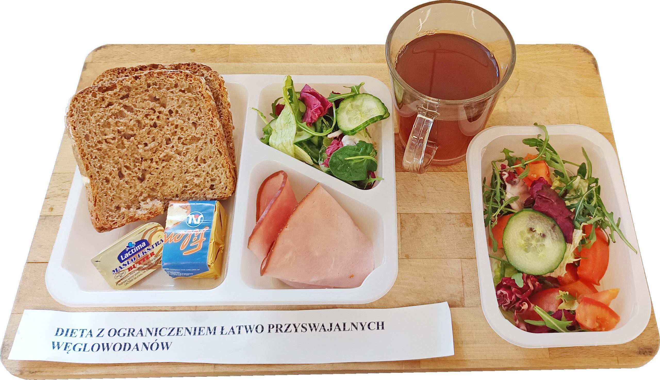 Śniadanie diety z ograniczeniem łatwo przyswajalnych węglowodanów: kromki chleba pełnoziarnistego, masło, plasterki wędliny drobiowej oraz świeże warzywa. Obok sałatka z mixu sałat, ogórka i pomidora oraz kubek herbaty