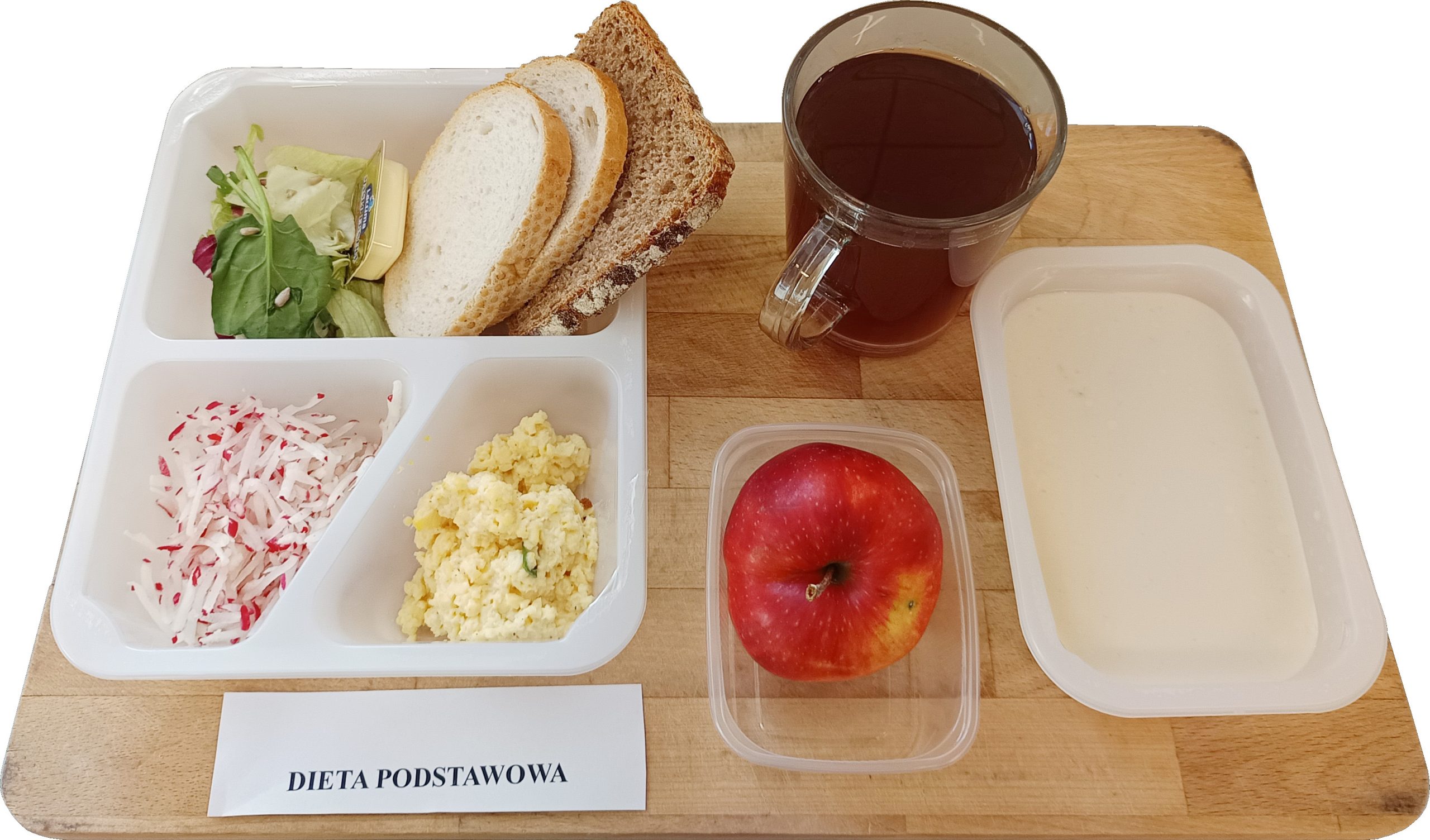 Na drewnianej tacy zestaw śniadaniowy diety podstawowej: kromki chleba jasnego i razowego, masło, mix sałat z pestkami, pasta jajeczna oraz surówka z rzodkiewki. Obok kubek herbaty, jabłko w pojemniku i jasna zupa w osobnym naczyniu