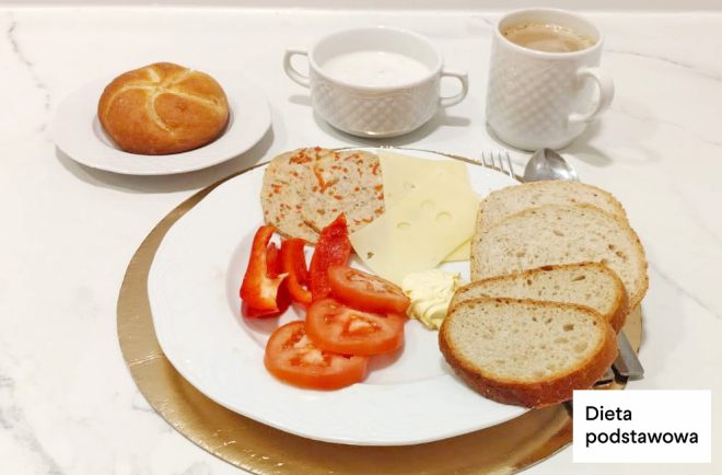 Śniadanie w diecie podstawowej: pieczywo pszenne, plaster sera, wędlina drobiowa, pomidor i papryka. W tle bułka, kubek kawy oraz napój mleczny
